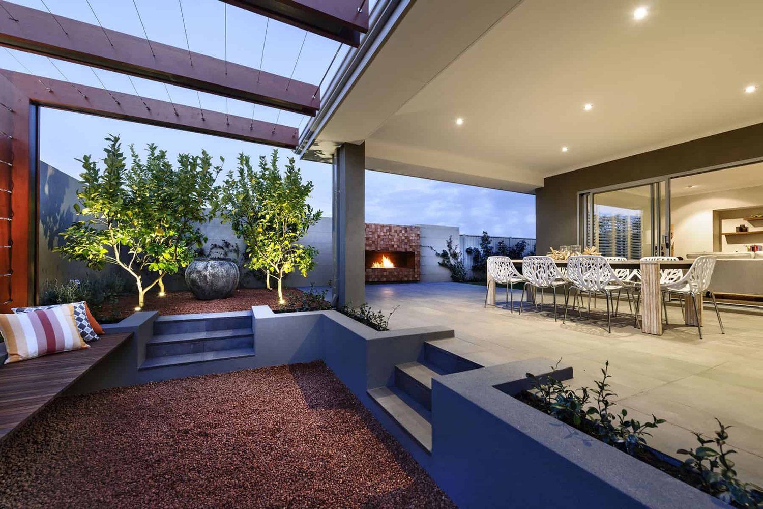 Alfresco dining and courtyard at dusk with a lit brick fireplace, citrus trees and a stone water urn